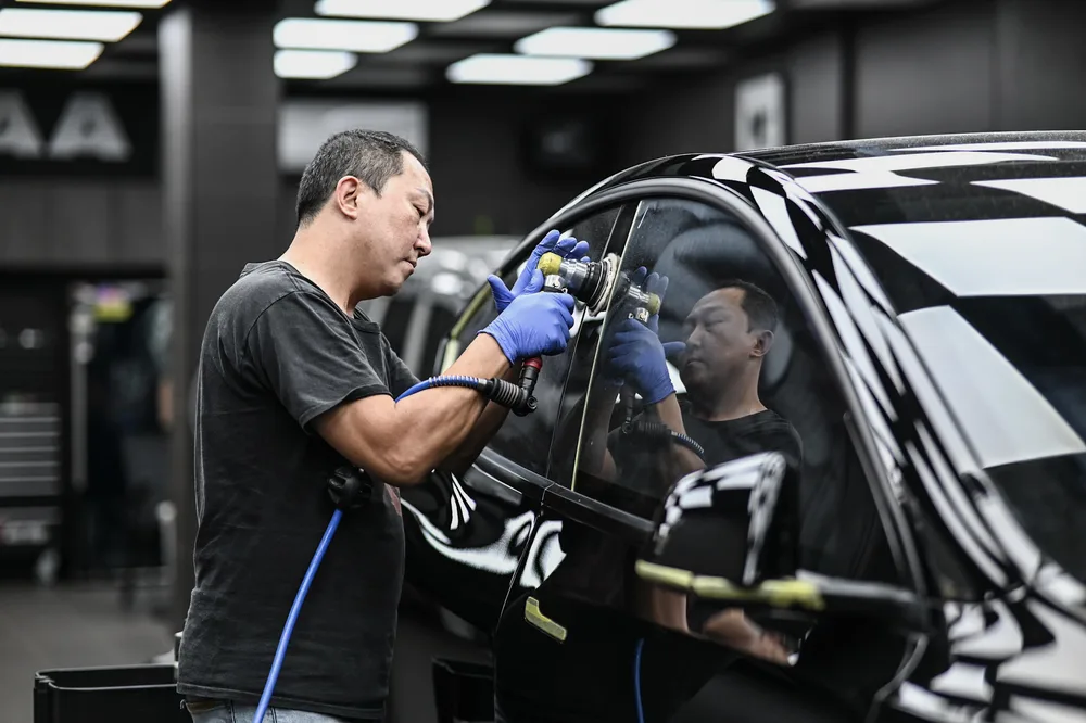 技師在黑色 Tesla 車側以研磨機進行玻璃拋光鍍膜，車窗映出施工區棋盤格天花板 誠美車體美容