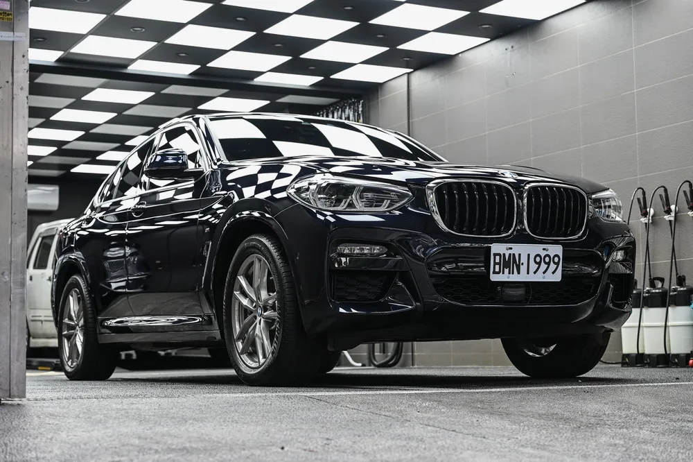 黑色 BMW X4 M Sport 完成基礎護理後停於格紋施工區，左前方全景鈑面映出天花板倒影，誠美車體美容